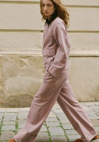 Różowy dwuczęściowy strój składający się z kurtki z kołnierzykiem z przednimi kieszeniami oraz luźnych spodni. Tkanina wydaje się miękka o gładkiej fakturze.