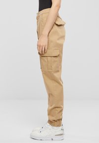 Beige cargo kalhoty s bočními kapsami, manžetovými kotníky a hladkým povrchem, doplněné bílými teniskami pro kontrast.