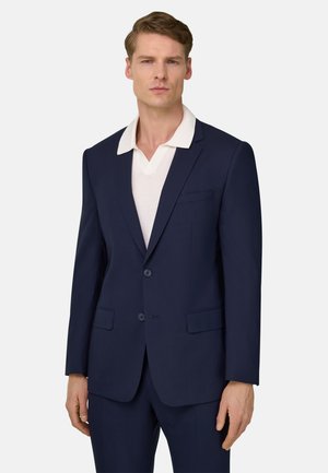 Uomo che indossa una giacca e pantaloni blu scuro con una camicia bianca con colletto, su uno sfondo chiaro uniforme.