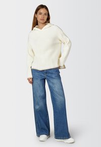 Hellbeige, gerippter Strickhoodie mit lockerer Passform, kombiniert mit weit geschnittenen blauen Jeans und weißen Sneakers. Schlichtes Design mit Kapuzendetail.