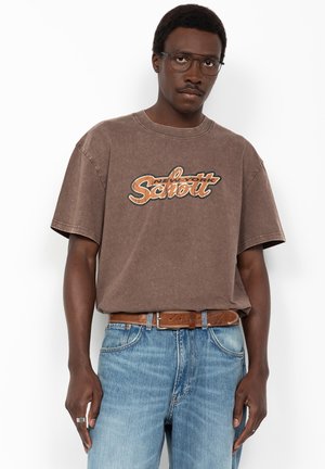 Mann trägt braunes "New York Scout" T-Shirt, blaue Jeans, braunen Gürtel und Brille, steht vor weißem Hintergrund.