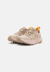 Beige idrettssko med en strukturert mesh-overdel, rosa gummisåle, og lyse gule detaljer på hælflappen og tungen.