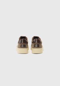 Tenisi din suede maro cu accente din piele maro la călcâi și tălpi din cauciuc crem. Au o etichetă cu logo pe călcâi și modele texturate pe talpă.