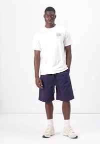 Hvit bomulls t-skjorte med trykket logo, kombinert med marineblå shorts. Antrekket fullføres med lyse sneakers og hvite sokker.
