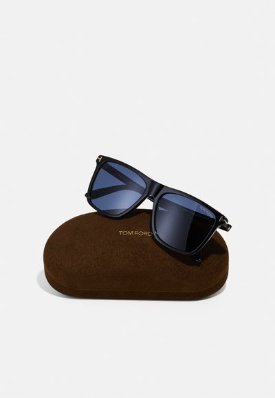 Occhiali da sole neri quadrati con lenti blu scuro, dettagli in oro e montatura spessa, appoggiati su una custodia in pelle scamosciata marrone con l'iscrizione "TOM FORD".