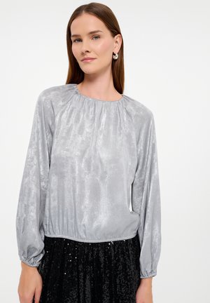 Zilveren blouse met lange mouwen, een gegatherde halslijn en een elastische zoom. Heeft een zachte, glanzende textuur en wordt gecombineerd met een zwarte rok met pailletten.