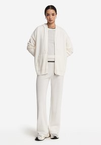 Vit hoodie med dragkedja fram, två fickor och avslappnad passform, tillsammans med matchande vita byxor. Båda är tillverkade av mjukt material.