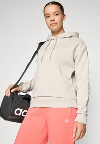 Beige hoodie met een voorzak en verstelbare koordjes. Roze sweatpants met een klein logo. Zwarte sporttas met witte branding.