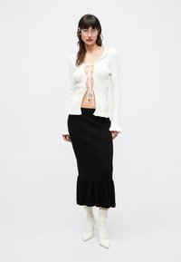 Valge pikakäise ja ees väikeste sõlmedega pluus, millel on volangidega mansetid, sobitatud musta soonikkoes midi seelikuga, millel on laiutuva äärega. Jalanõudeks on valged terava ninaga kontsakingad.