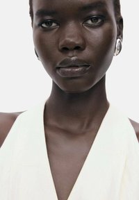 Haut blanc avec un décolleté en V profond. La peau semble lisse et foncée, encadrée par des cheveux courts. Les traits incluent des lèvres bien définies et des boucles d'oreilles en argent.