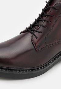 Bota de vestir de cuero marrón con acabado suave, punta redonda, cordones negros y suela de goma con detalles texturizados a lo largo del borde.