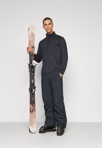 Schwarzes Quarter-Zip-Oberteil, dunkle Schneehosen, hält einen Ski mit einem beige-rosa Design. Schwarze Skischuhe mit grünen Akzenten. Neutraler Hintergrund.