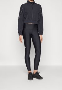 Černá krátká bunda s vysokým límcem a elastickými manžetami, ve spojení s těsně přiléhavými černými legínami. Oba kousky jsou vyrobeny z elastické, hladké látky.