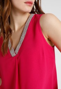 Fuchsia ärmlös topp med V-ringning prydd med silverfärgad pärlkant, tillverkad av ett mjukt, lätt tyg med en slät textur.