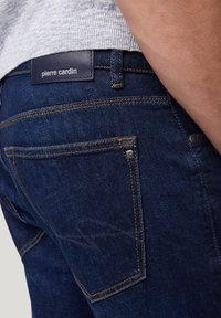Donkerblauwe denim jeans met oranje stiksels, stikseldetail op de achterzak en een zwart leren merklabel "pierre cardin" op de tailleband.