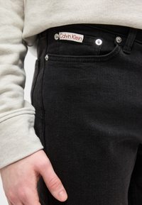 Άτομο που φοράει μαύρο τζιν Calvin Klein και μπεζ μακρυμάνικη μπλούζα, το χέρι χαλαρό δίπλα στο μπροστινό τσέπη.