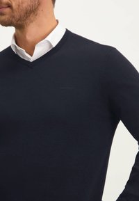 Marineblauwe gebreide trui met een V-halsontwerp, voorzien van een zachte textuur en subtiele branding nabij de borst. Lange mouwen met ribgebreide manchetten.