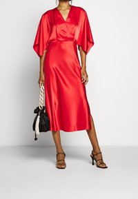 Robe en satin rouge avec un décolleté en V et de larges manches style cape. Présente une coupe mi-longue et une fente latérale, associée à un sac noir et des talons.