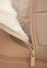 Beige schoen met een zachte, crèmekleurige bontvoering en ritsdetails. Het materiaal is een combinatie van gladde en gestructureerde oppervlakken. De rubberen zool is zichtbaar.
