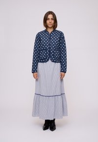 Donna in piedi, che indossa una giacca blu con motivo, una lunga gonna a righe azzurro chiaro con orlo a balze e stivali neri alla caviglia su uno sfondo uniforme.
