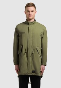 Homme portant une longue veste vert olive avec un col haut, une fermeture éclair frontale, des poches latérales avec rabats et des cordons ajustables à la taille et à l'ourlet.