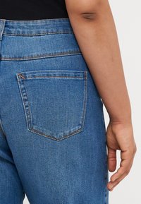 Οι μπλε τζιν παντελόνες διαθέτουν μεσαίο ύψος, με μια μόνο πίσω τσέπη και αντίθετες ραφές. Ο ιστός φαίνεται λείας και υφής.