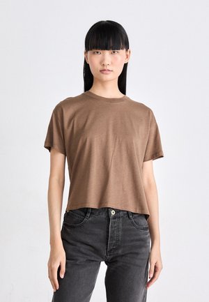 Jonge vrouw met lang zwart haar, gekleed in een bruin shirt met korte mouwen en zwarte high-waisted jeans, staand tegen een witte achtergrond.