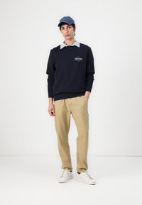 Marine sweatshirt met logo, gelaagd over een wit gestreept shirt, gecombineerd met kaki broeken en witte sneakers, afgewerkt met een blauwe pet.