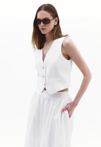 Weißes ärmelloses Oberteil mit goldenen Knöpfen und passendem Rock mit elastischem Bund. Glatte Textur mit einer taillierten Silhouette.