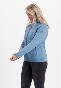Ljusblå zip-up jacka med en vadderad textur, ståkrage och åtsittande ärmar. Kombineras med mörka jeans.