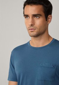 Modré tričko s krátkým rukávem vyrobené z bavlny, s kulatým výstřihem a kapsou na levé straně hrudníku, která má jemné švy.