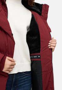Bordo pūkuotas švarkas su juodu dirbtiniu kailiu, užsegamas užtrauktuku ir šoninėmis knopkėmis, dėvimas ant balto megztinio ir džinsų.