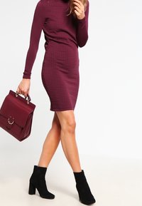 Robe bordeaux à manches longues avec motif texturé, silhouette ajustée, assortie à un sac à main et des bottines noires.