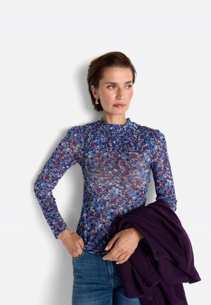 Woman wearing a blue and purple floral long-sleeve top, holding a dark purple coat, with one hand in her jeans pocket.