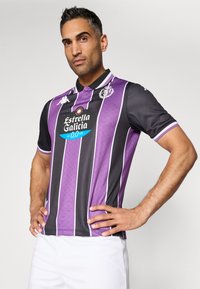 Camisa de fútbol a rayas moradas y negras con cuello, que presenta un patrón texturizado, mangas cortas y un gran logo en la parte delantera.