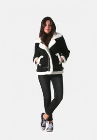 Veste en peau lainée noire avec col en polaire blanche, dotée d'une fermeture éclair à l'avant, de poches latérales et d'accents noirs. Portée avec un jean skinny noir et des baskets.