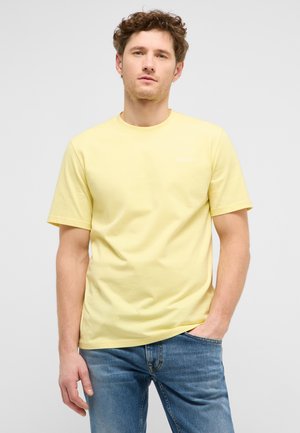 Young man with curly hair wearing a plain yellow short-sleeve t-shirt and blue jeans, standing with one hand in pocket.