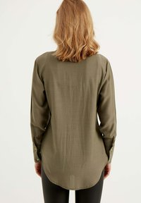 Olive green koszula z długim rękawem o zaokrąglonym dołem, wykonana z gładkiego materiału, z zapięciem na guziki na mankietach oraz detalem na karku.