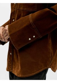 Brun skjorte i semsket skinn med avslappet passform, store mansjetter, kontrastsøm og trykknapper på ermene. Teksturert finish synlig.