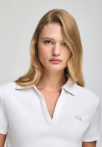 Camiseta polo blanca de manga corta y escote en V, con un sutil logo "POLO CLUB" en gris en la parte izquierda del pecho. Textura de tela suave.