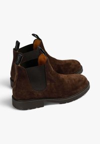 Botines de ante marrón con paneles laterales elásticos y lengüetas, que presentan un acabado texturizado y suelas de goma con un ligero patrón de rodadura.
