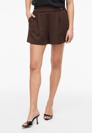 Kvinne iført mørkebrune shorts med høyt liv og knapper og svarte åpne høye hæler, står med en hånd i lomma.