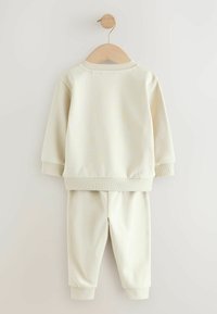 Conjunto de sudadera y pantalones para niños en color beige claro. Material suave, puños y dobladillo acanalados, ajuste relajado, colgado en una percha de madera sobre un fondo claro.