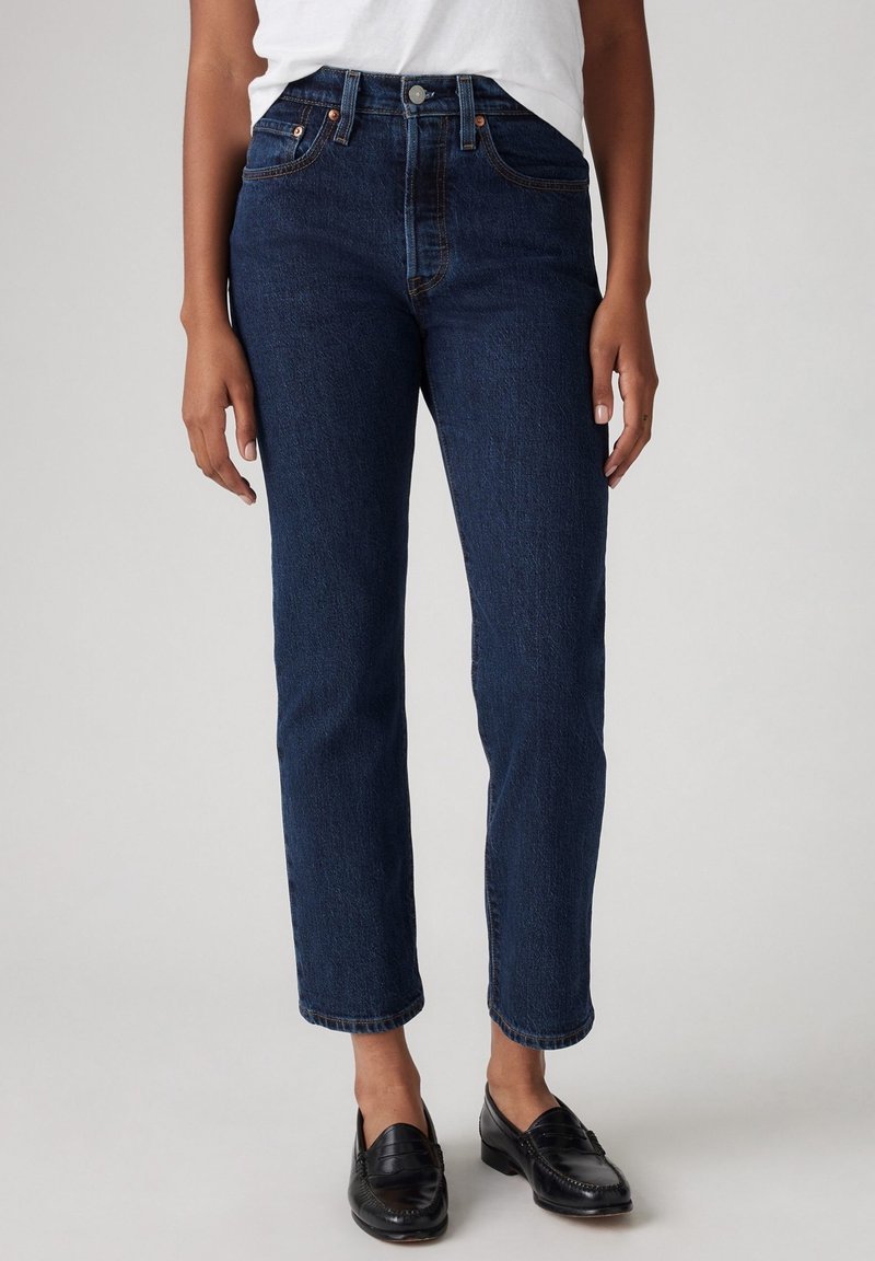 Personne portant un jean droit bleu foncé avec un haut blanc et des mocassins noirs, debout devant un fond gris uni.