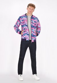 Giacca bomber colorata con un motivo camouflage nei colori rosa, blu e viola, con polsini a coste e zip frontale, indossata sopra una maglietta grigia.