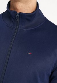 Navy zip-up jacket featuring a high collar, smooth fabric, and a small logo accent in red, white, and blue on the left chest area.