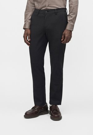Homme portant un pantalon noir sur mesure, des chaussures marron habillées et une chemise taupe à manches longues, debout sur un fond blanc uni.