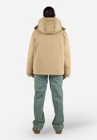 Beige Pufferjacke mit großer Kapuze, aus leichtem Material. Sie hat eine glatte Textur und ein Design mit überschnittenen Schultern und wird über türkisfarbenen Jeans getragen.