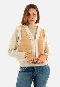 Gilet en daim beige avec des bords en fausse fourrure blanche, design en V et sans manches. Porté sur un pull léger et un jean bleu.
