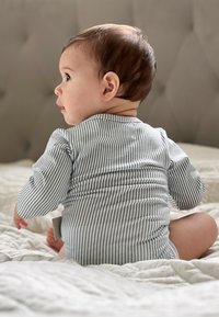 Gestreifter Baumwoll-Overall in Blau und Weiß, mit langen Ärmeln und einem Knopfdetail auf der Rückseite, getragen von einem sitzenden Baby auf einer hellen Decke.